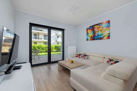 A spacious lounge view highlights the open-concept layout connecting the seating area to the garden. A large flat-screen TV is mounted on the wall across from the comfortable sectional sofa.