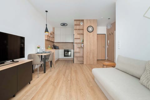 A bright living room features a comfortable grey sofa, wooden flooring, and a sleek TV stand. The open-plan layout connects the lounge area with a modern kitchenette in the background.