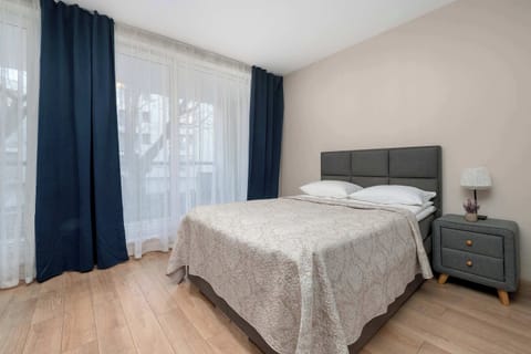 A minimalist-style apartment features white walls, warm wooden flooring, a light gray bedspread, blue curtains, a single grey bedside table, a modern wall light, and a small table with two matching gray chairs.