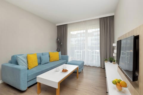 A light blue sofa is accented with bright yellow pillows and sits across from a long white media console. Large glass doors leading to a balcony allow plenty of natural light to fill the living room.
