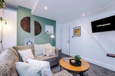 Cozy living room with a patterned sectional sofa, soft cushions, and a round wooden coffee table. Features a green accent wall with modern decor, wall-mounted TV, and warm lighting—perfect for relaxing or streaming your favorite shows in comfort.