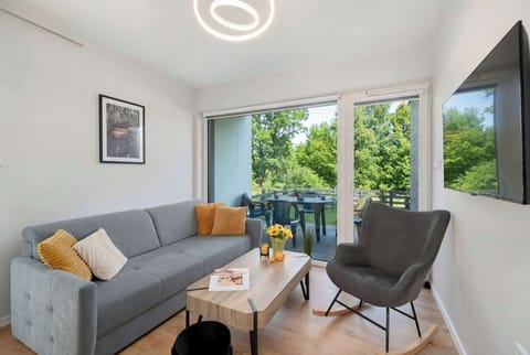 A cozy living room with a gray corner sofa and a modern armchair positioned near large windows. A small coffee table stands in the center of the seating area. Large windows bring in plenty of natural light and offer a view of greenery outside.
