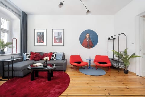 A spacious living room features a dark grey corner sofa adorned with vibrant red and patterned pillows, centered around a black coffee table on a plush red rug. A unique circular portrait hangs on the white wall, complemented by warm wooden flooring 