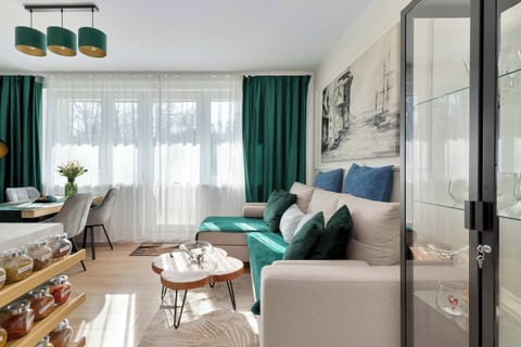 A stylish living room corner with a couch, coffee table, and modern decor. Green curtains and soft neutral colors give the space a calm and inviting feel. Natural light enters through a large window beside the seating area.