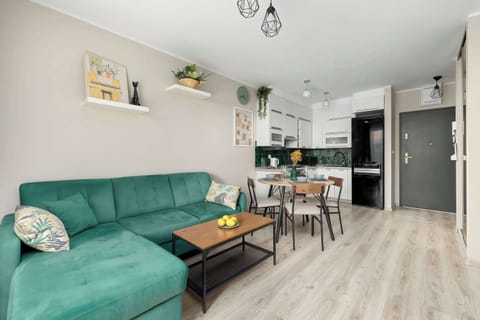 A spacious living room filled with natural light features a comfortable, dark teal sectional sofa on the left and a view of a dining area with a round table and a small kitchenette at the back.