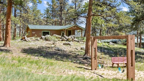 Outdoor play area with wooden swing set, red bench, and grassy yard framed by pine trees.