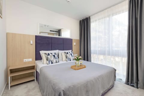 A large bed features a light grey duvet and matching pillows against a warm wooden headboard. Natural light flows into the room from a nearby window, highlighting the clean and modern design.