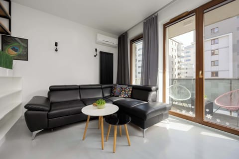 A bright living room with a dark sofa placed near large glass balcony doors. Natural light fills the room through tall windows with curtains. A small coffee table sits in front of the sofa, creating a cozy sitting area.