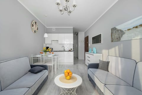 A brightly lit apartment features a gray sofa on one side and a dining table with chairs next to a white kitchen counter on the other. Yellow flowers and other small decorative items are visible in the background.