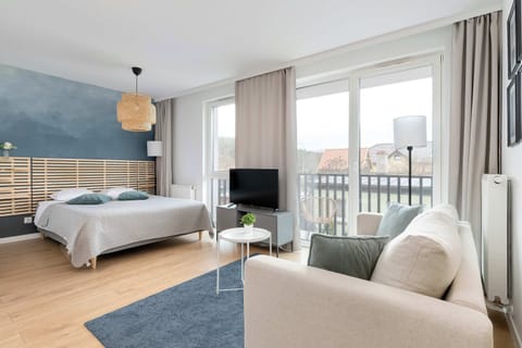A bright studio-style living and sleeping area with a bed near large windows, a small sofa, a TV stand, and a balcony visible outside. Natural light fills the modern space with light wood flooring and neutral decor.