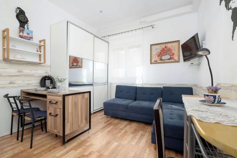 A studio apartment features a built-in desk with two chairs, a blue corner sofa, and a kitchen unit with upper shelves. The space is compact and utilizes wood-tone flooring and light furniture to create an airy feel.