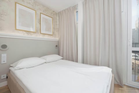 Soft natural light filters through floor-to-ceiling curtains next to a large, comfortable bed. The headboard area is illuminated by subtle integrated lighting, highlighting the elegant wall textures.