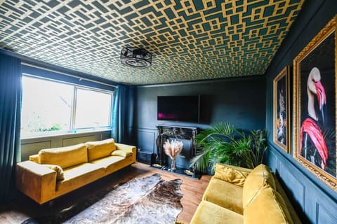 The living room features dark-painted walls, a bold gold-and-teal patterned ceiling, two yellow velvet sofas, an ornate fireplace surround, large tropical plants, and a wall-mounted TV.