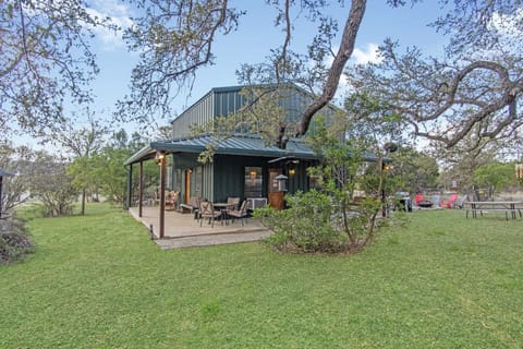 A side view of the forest-green metal cottage reveals an expansive wrap-around porch with outdoor seating, nestled under the shade of mature oak trees and overlooking a spacious lawn with a fire pit and picnic area.