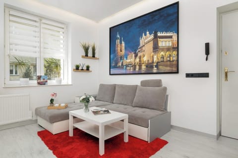 A modern living room with a gray corner sofa and a contrasting red rug under a white coffee table. The wall is dominated by a large-format print depicting Kraków’s Main Square at night.