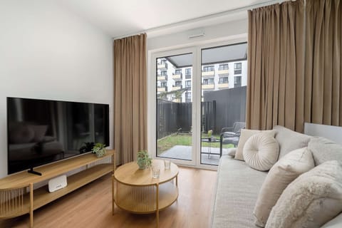 A sun-drenched living area features a plush beige sectional sofa and a sleek wooden TV stand. Large glass doors open directly onto a private garden patch, creating a seamless connection to the outdoors.