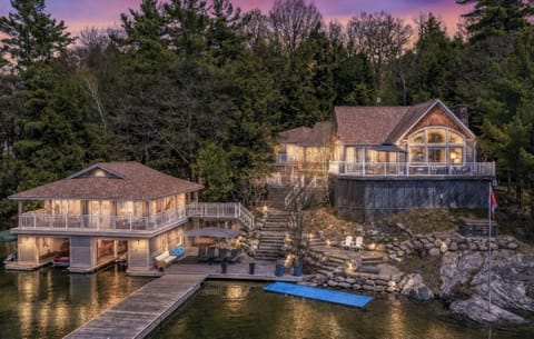 Lakefront view of the boathouse with wraparound balcony, dock, stone steps, and adjacent main cottage at dusk.