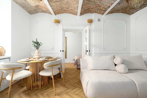 A bright dining space features a slatted wooden table surrounded by white chairs under a stunning vaulted brick ceiling. The room flows into a minimalist living area with a plush beige sofa and ornate wall molding.