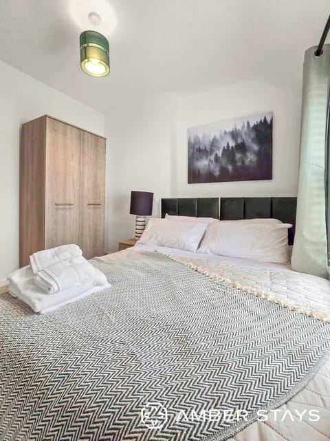 A bright, modern bedroom featuring monochrome patterned bedding, a wooden wardrobe, and forest wall art. The space is accented by a green pendant light and a sleek bedside lamp.
