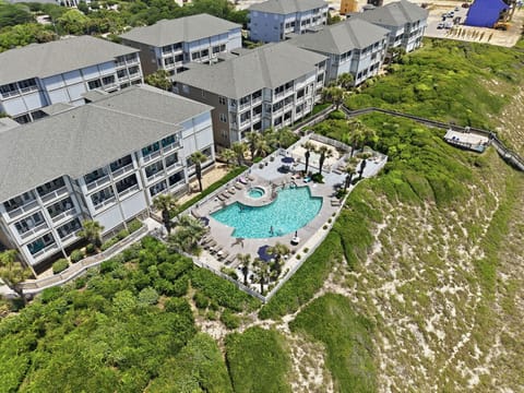 Aerial view of the vacation rental property featuring multi-story buildings surrounding a central pool area with hot tub, bordered by natural coastal landscape and walkways.