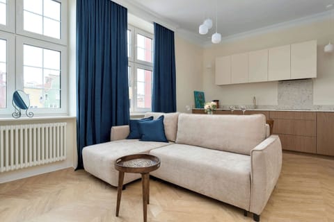 A bright living room features a light-colored sofa, a wooden coffee table, and large windows with deep navy blue curtains. The open layout leads into a minimalist kitchenette area with wood-toned cabinetry.