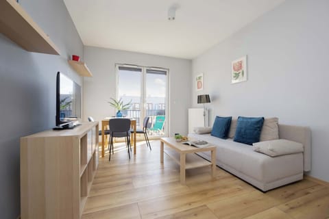 Light-filled living room features a minimalist wooden media console and a comfortable grey sectional sofa. Polished wood flooring runs throughout the space, leading toward a bright balcony door.