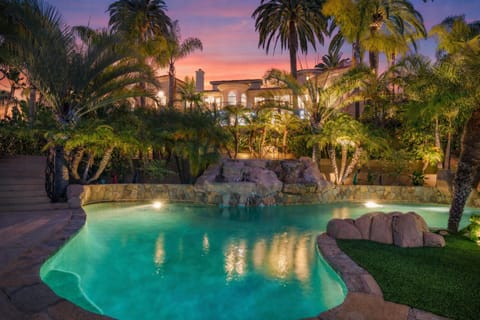 Resort‑style backyard with a glowing pool, lush palms, and a grand estate backdrop.