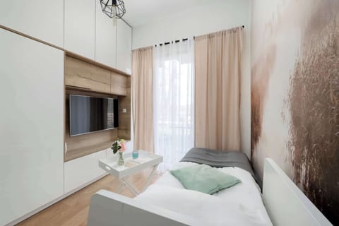 A minimalist studio interior features a single bed positioned against a scenic landscape wall mural. The room is equipped with white cabinetry and large windows draped in neutral beige curtains.
