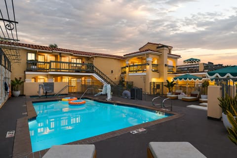 Resort-style pool courtyard with lounge seating, glowing Spanish-inspired architecture, and a sunset sky.