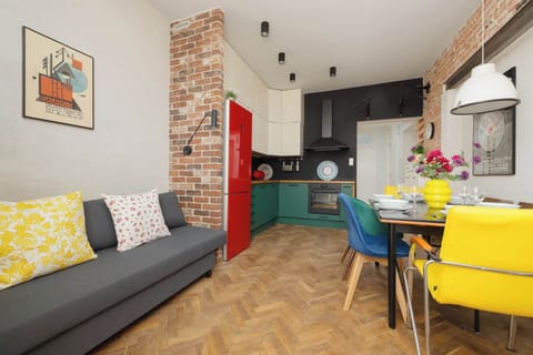 A bright and open living area features a grey sofa  and a dining table set for four near the balcony entrance. The room is illuminated by natural light and decorated with yellow and patterned accent pillows.