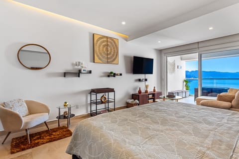 Spacious bedroom layout featuring a large bed with patterned linens, wall-mounted TV, and glass doors opening to a coastal balcony.