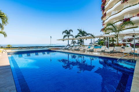 Large blue-tiled swimming pool surrounded by white lounge chairs, umbrellas, palm trees, and the resort building.