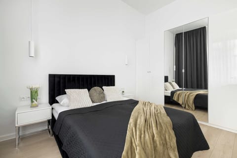 A bright and minimalist bedroom features a large bed with a dark duvet and a decorative gold throw. A tall, dark mirror stands in the corner, reflecting the clean, white walls of the room.