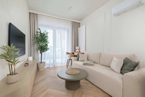 A bright living room features a textured beige sofa, a minimalist olive-green coffee table, and chevron wood flooring. Large glass doors leading to a balcony fill the space with natural light.