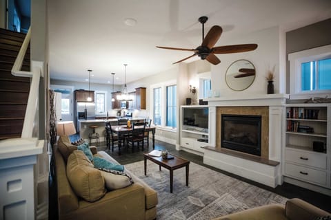 Welcome to Riverwalk Path!  A wide view of the open-concept main level showcasing the living area, gas fireplace with built-in shelving, and the kitchen beyond, all connected under warm ceiling fan lighting.