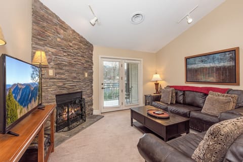 Relax in your cozy living room with stone fireplace, plush leather seating, and mountain views through glass doors to your private balcony.