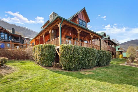 Rustic log cabin with wraparound porch, stone chimney, green roof, and hills