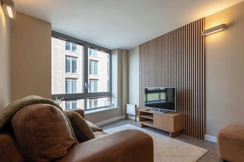 Bright and modern living room with large windows that fill the space with natural light and city views. Features a cozy leather sofa, sleek wooden accents, and a minimalist TV setup for a relaxing stay.