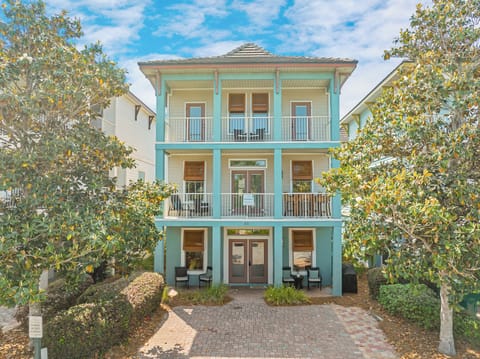 A charming three-story coastal retreat with turquoise façade, private balconies, and lush garden landscaping framed by mature trees.