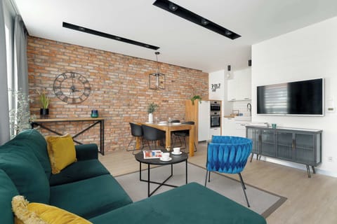 A spacious living room is furnished with a comfortable green sectional sofa and a teal accent chair arranged around a coffee table, set against a stylish exposed brick wall. A mounted TV and storage console are visible on the adjacent white wall.