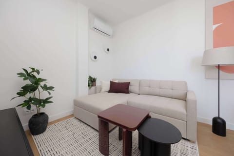 A modern living area with a light-colored sofa placed against the wall and a compact dining table with two stools in the center. A tall plant adds a natural touch, while the wall-mounted air conditioning unit ensures comfort. 