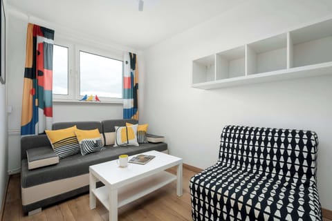 A cozy living area with a patterned sofa, a small coffee table, and a TV setup. Colorful accents on the cushions and curtains bring life to the otherwise neutral palette. The layout feels compact but comfortable for relaxing.