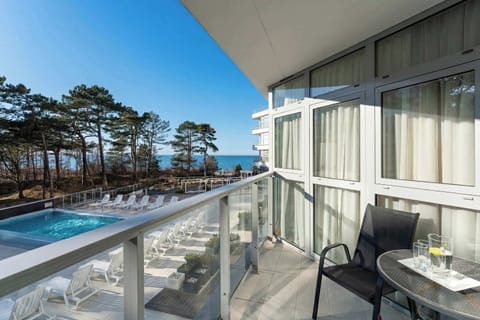 A balcony with a glass railing overlooking a swimming pool and surrounding greenery. Outdoor seating provides a perfect spot to relax and enjoy the view. The setting feels peaceful and ideal for unwinding in fresh air.