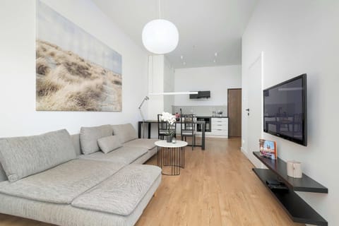 A brightly lit living room with light wood floors, a sofa, and a wall-mounted TV. A small dining area can be seen in the background.