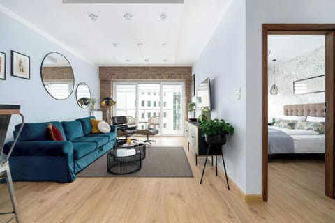 A spacious living room featuring a plush teal sofa, wooden flooring, and a large doorway leading to a bright bedroom.
