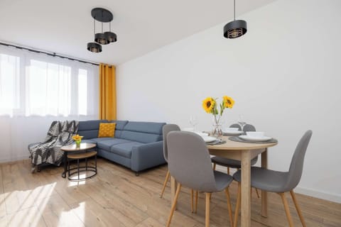 A bright living room with a grey sofa and a small dining table. Large windows with yellow curtains bring in plenty of natural light.