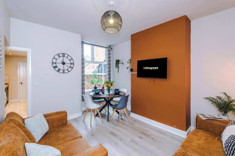 - Modern comfort meets elegance: tan leather sofas, patterned cushions, a burnt orange wall with mounted TV, framed art, and a round dining table set with wine and blue plates, surrounded by greenery and warm décor.
