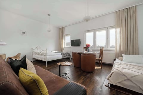 A wide-angle shot of an open-plan living and dining area, featuring dark wood floors, a sofa with a yellow pillow on the left, a wooden dining table with chairs, and windows with curtains.