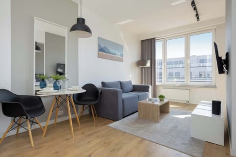 A sunlit studio apartment showing a modern living and dining area. It features a grey sofa, a wooden dining table with black chairs, and a large window overlooking trees.