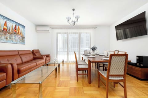 A spacious living room features a large, cognac-colored leather sofa and a wooden dining table with six chairs. Sunlight streams through large balcony windows, illuminating the polished wooden floors.